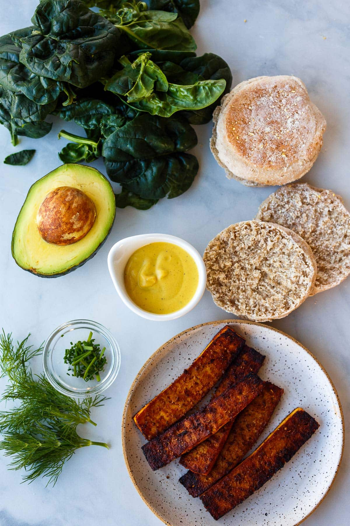 ingredients for vegan eggs benedict- english muffins, spinach, avocado, vegan hollandaise, tofu bacon, chives, dill.