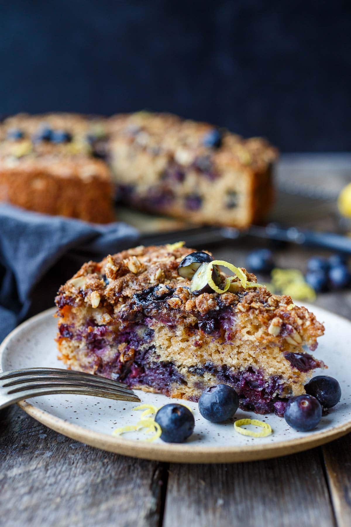 slice of blueberry coffee cake on plate with fresh blueberries and lemon zest.
