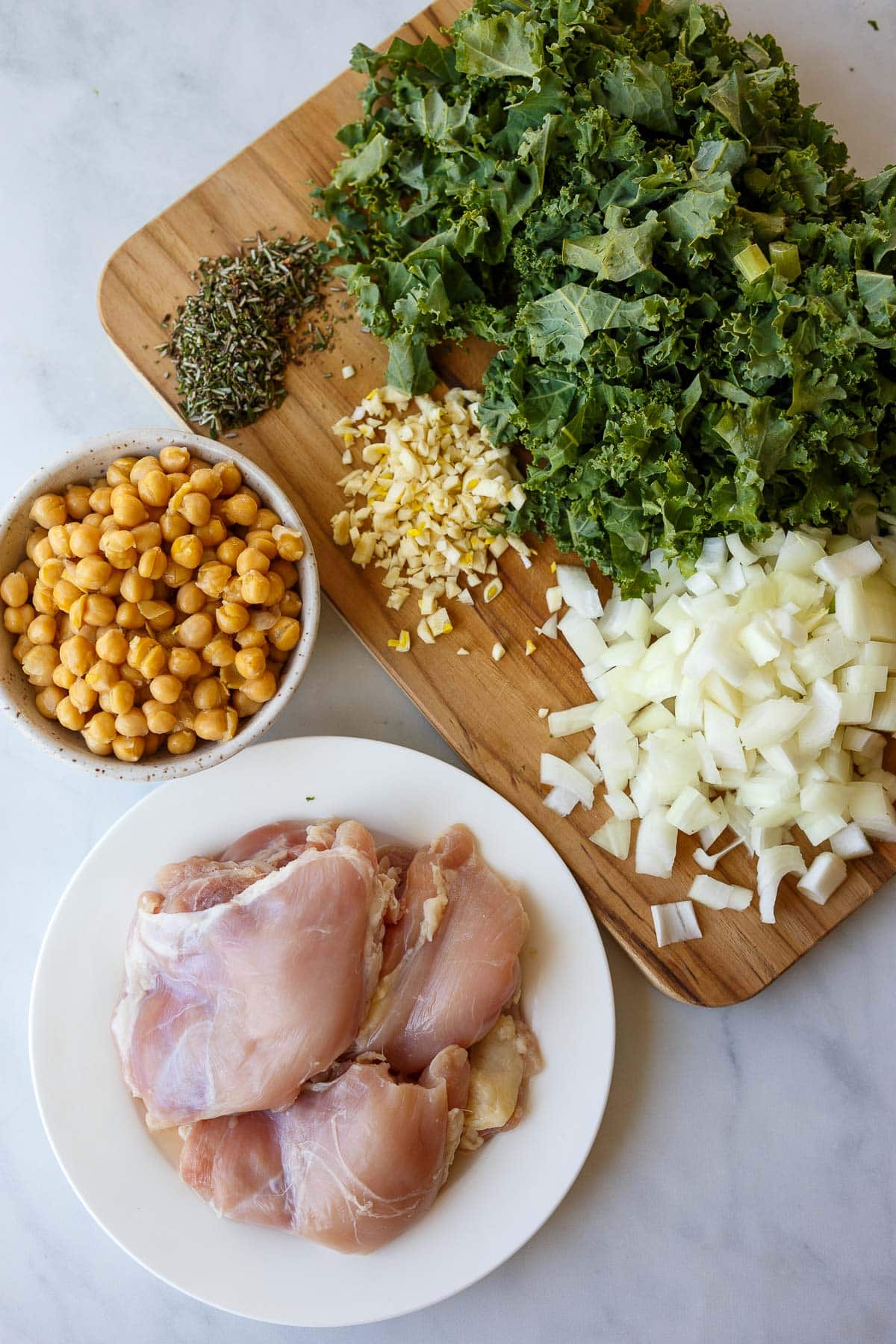 ingredients for chicken and kale soup - chopped kale, onion, garlic, rosemary, bowl of chickpeas, plate of chicken thighs. 