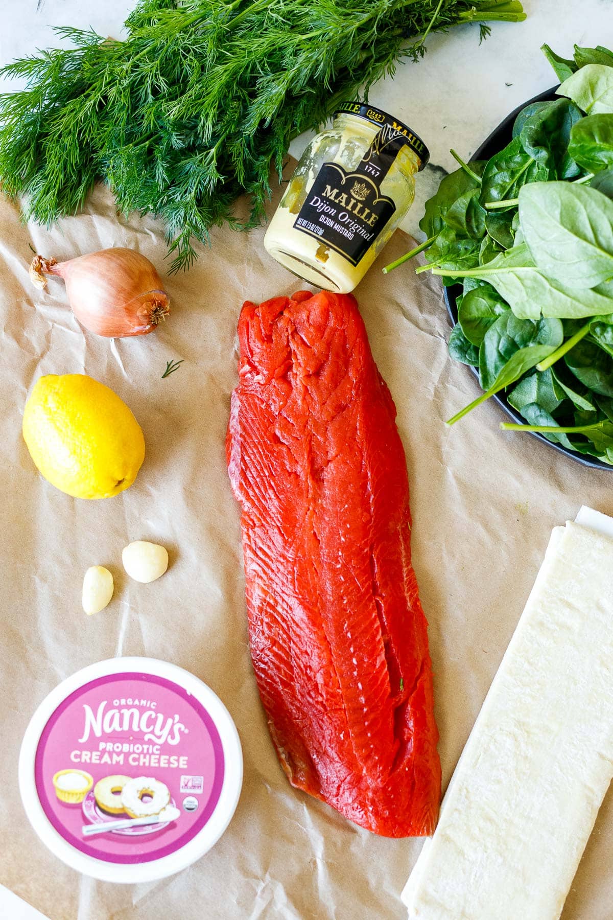 ingredients for salmon wellington- fresh dill and spinach, dijon, shallot, lemon, salmon, garlic, cream cheese, puff pastry, all laid out on parchment.