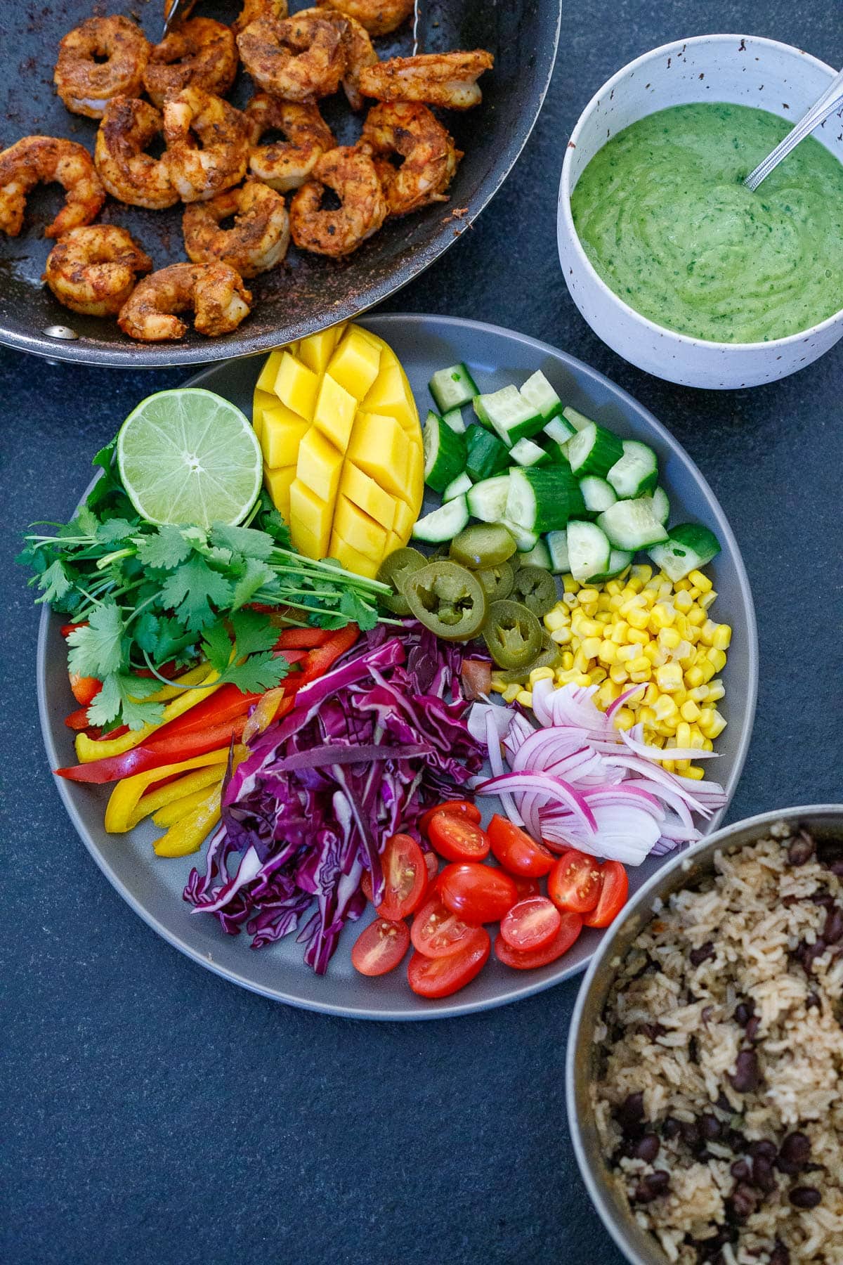 ingredients for spicy mexican shrimp and rice bowls - skillet with shrimp, creamy avocado sauce, plate with chopped and sliced veggies, mango, and herbs, and pot with rice and beans. 