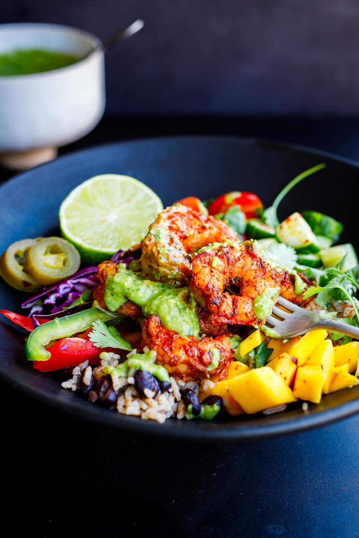 spicy Mexican shrimp served on rice and bean bowl with mango, bell peppers, cabbage, cucumbers, jalapeno, and lime, with creamy avocado sauce drizzled over top.
