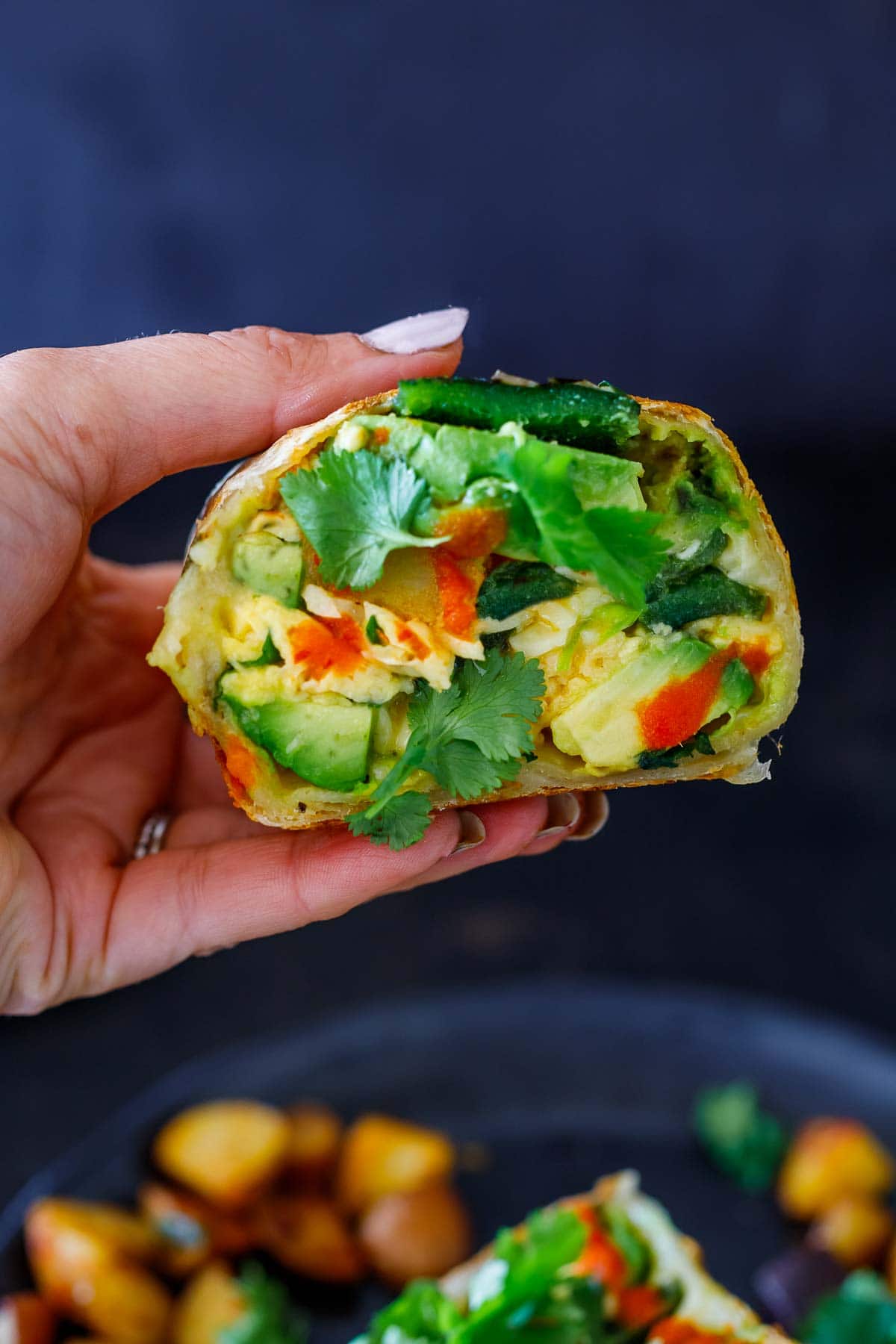 hand holding vegetarian breakfast burrito cut in half to reveal interior with eggs, cheese, avocado, poblano, cilantro, hot sauce.