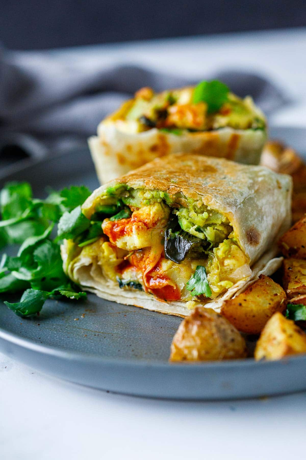 vegetarian breakfast burritos cut in half to reveal avocado, eggs, poblano, cilantro, and hot sauce- served on plate with potatoes.