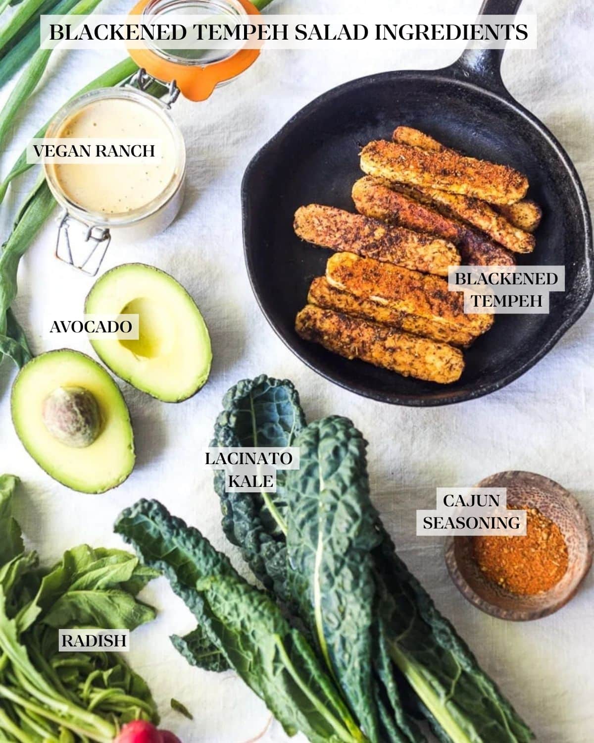 Blackened tempeh salad ingredients laid out and labeled - vegan ranch, avocado, blackened tempeh in a cast iron skillet, lacinato kale, cajun seasoning, and radish.