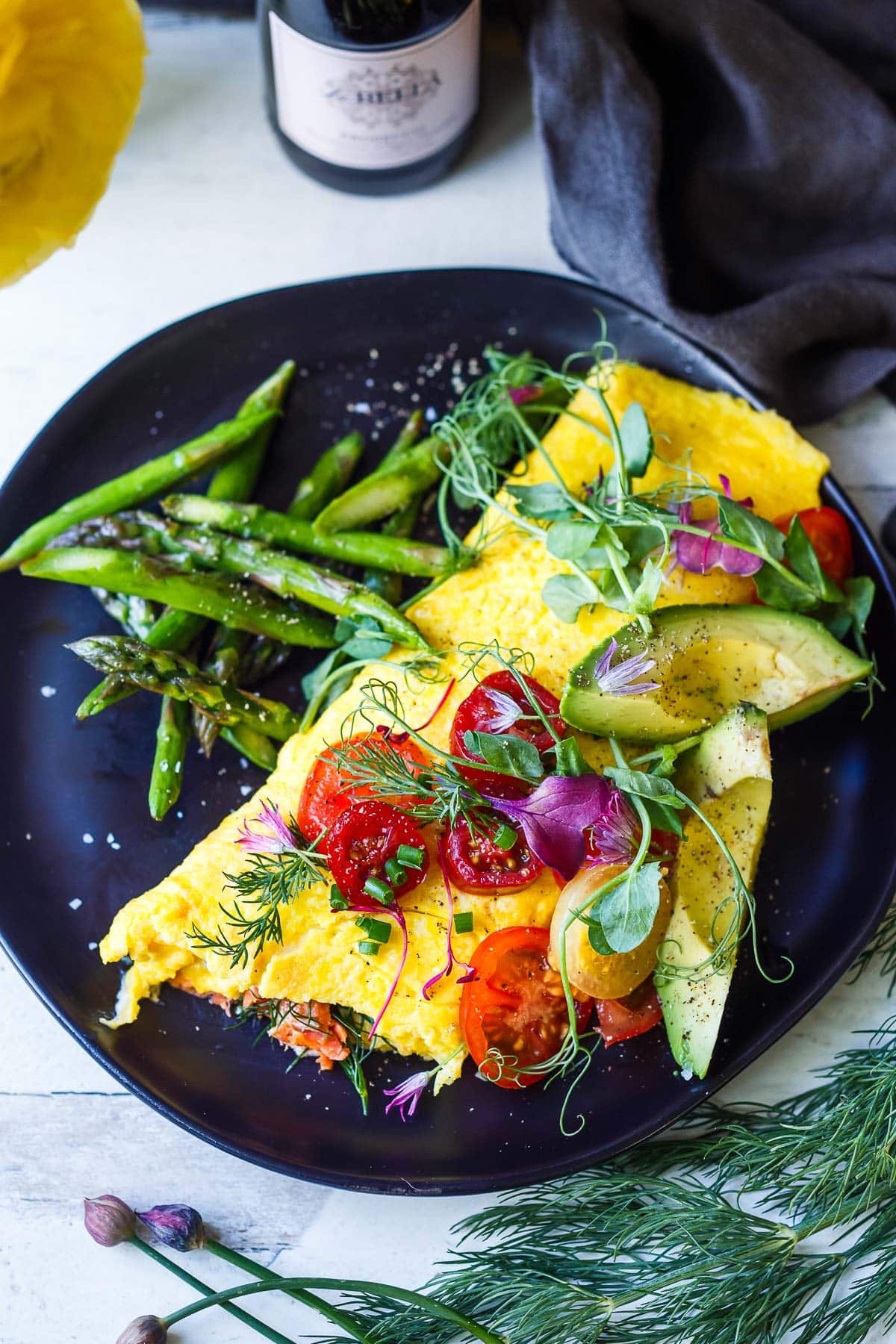 champagne omelette on plate with asparagus, topped with avocado slices, cherry tomatoes, micro greens, and herbs.