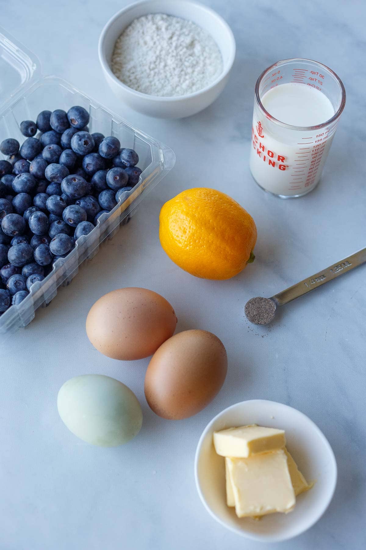 ingredients for blueberry dutch baby laid out- flour, milk, blueberries, lemon, eggs, cardamom, and butter.