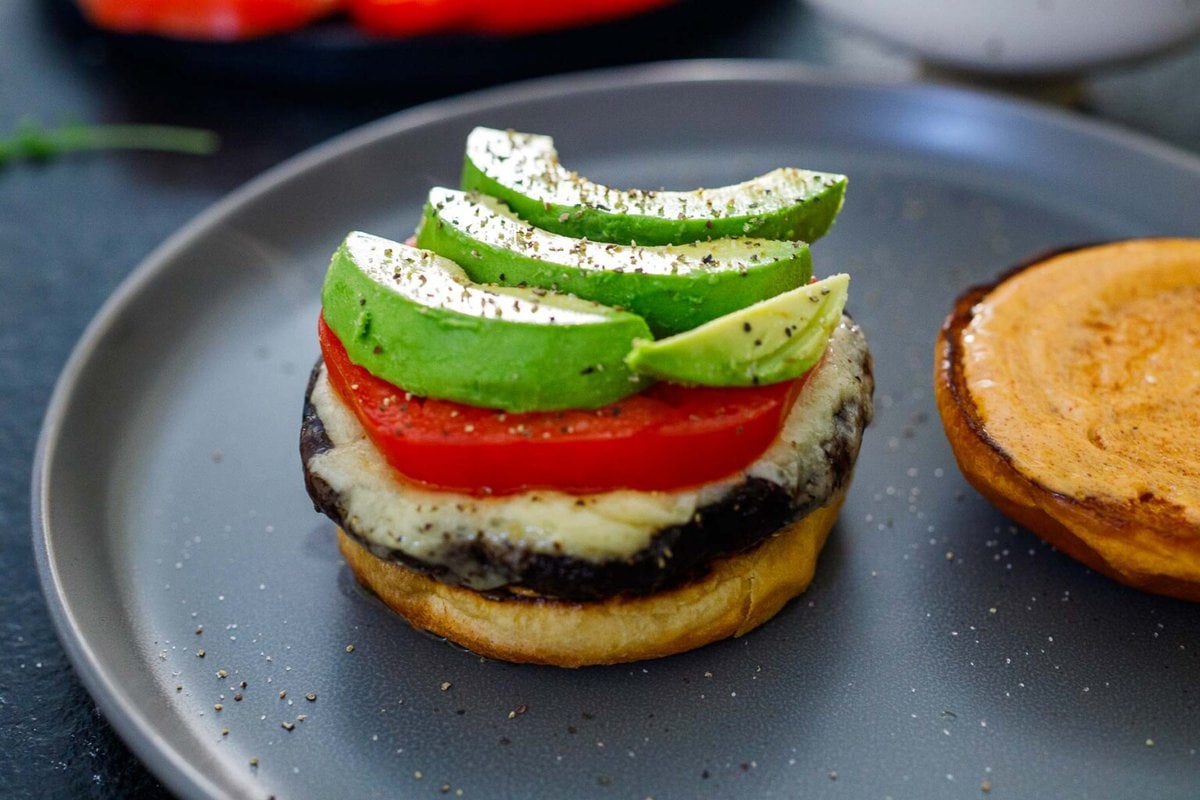 assembling portobello mushroom burger with brioche bun, stacked with roasted portobello, melted cheese, sliced tomato and avocado.