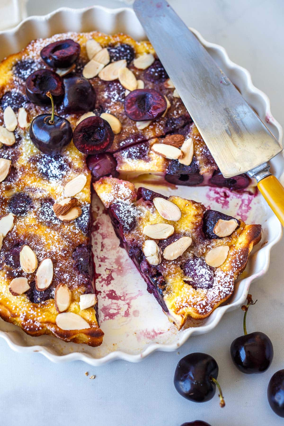 slices of cherry clafoutis in round baking dish, garnished with almond slices and powdered sugar.
