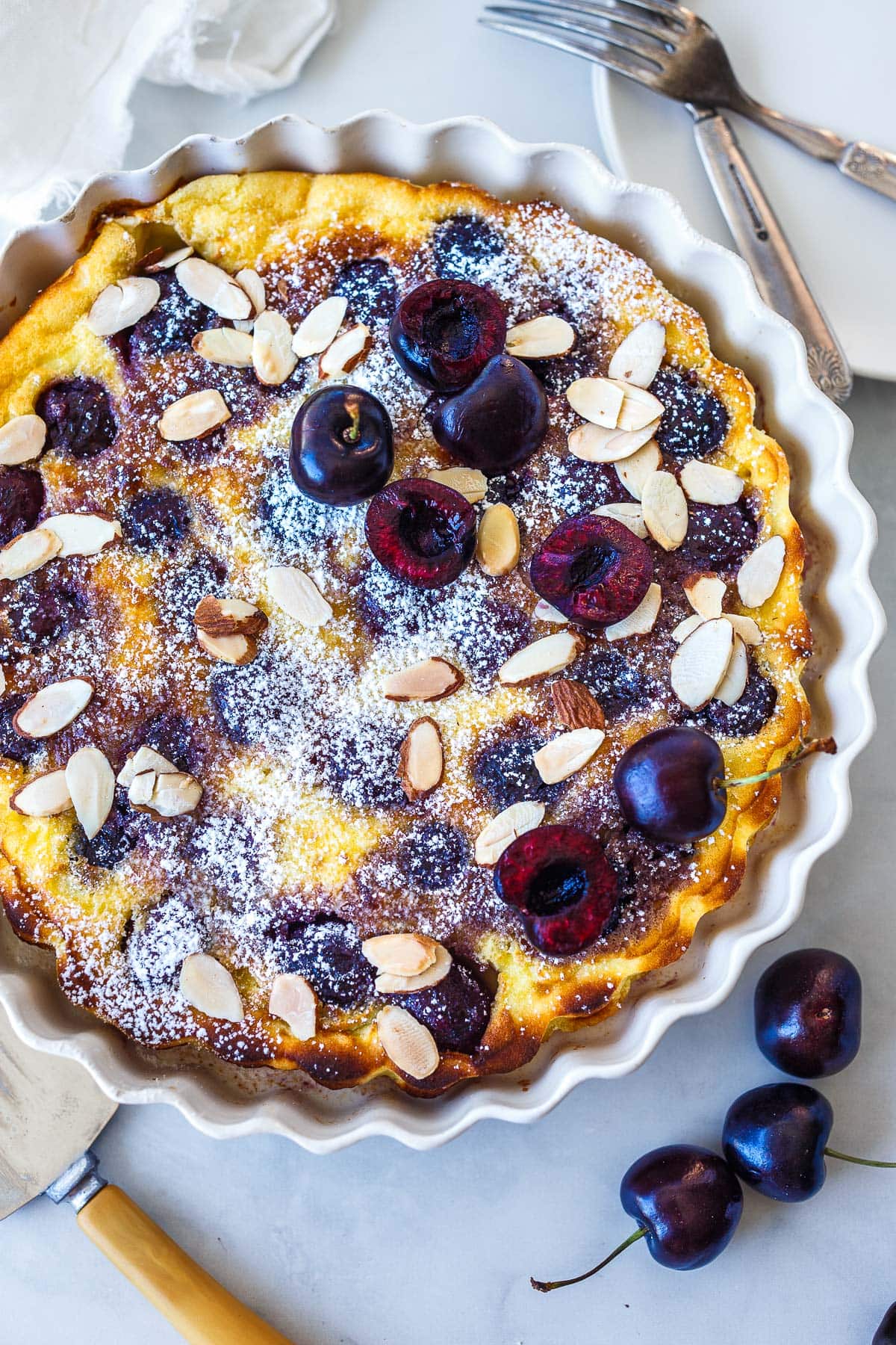 cherry clafoutis in round baking dish topped with sliced almonds and powdered sugar.