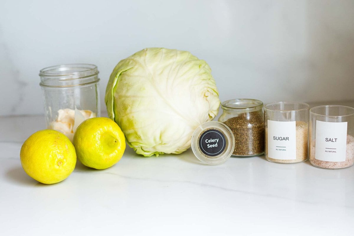 ingredients laid out for creamy coleslaw- mayonnaise, lemons, green cabbage, celery seed, sugar, and salt. 