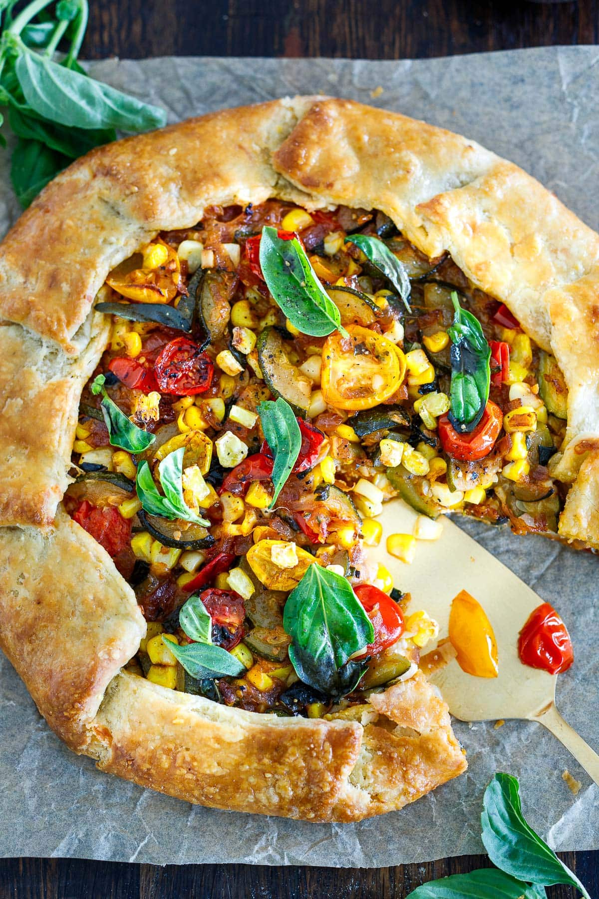 savory summer galette on parchment with fresh basil and piece missing with pie serving utensil.
