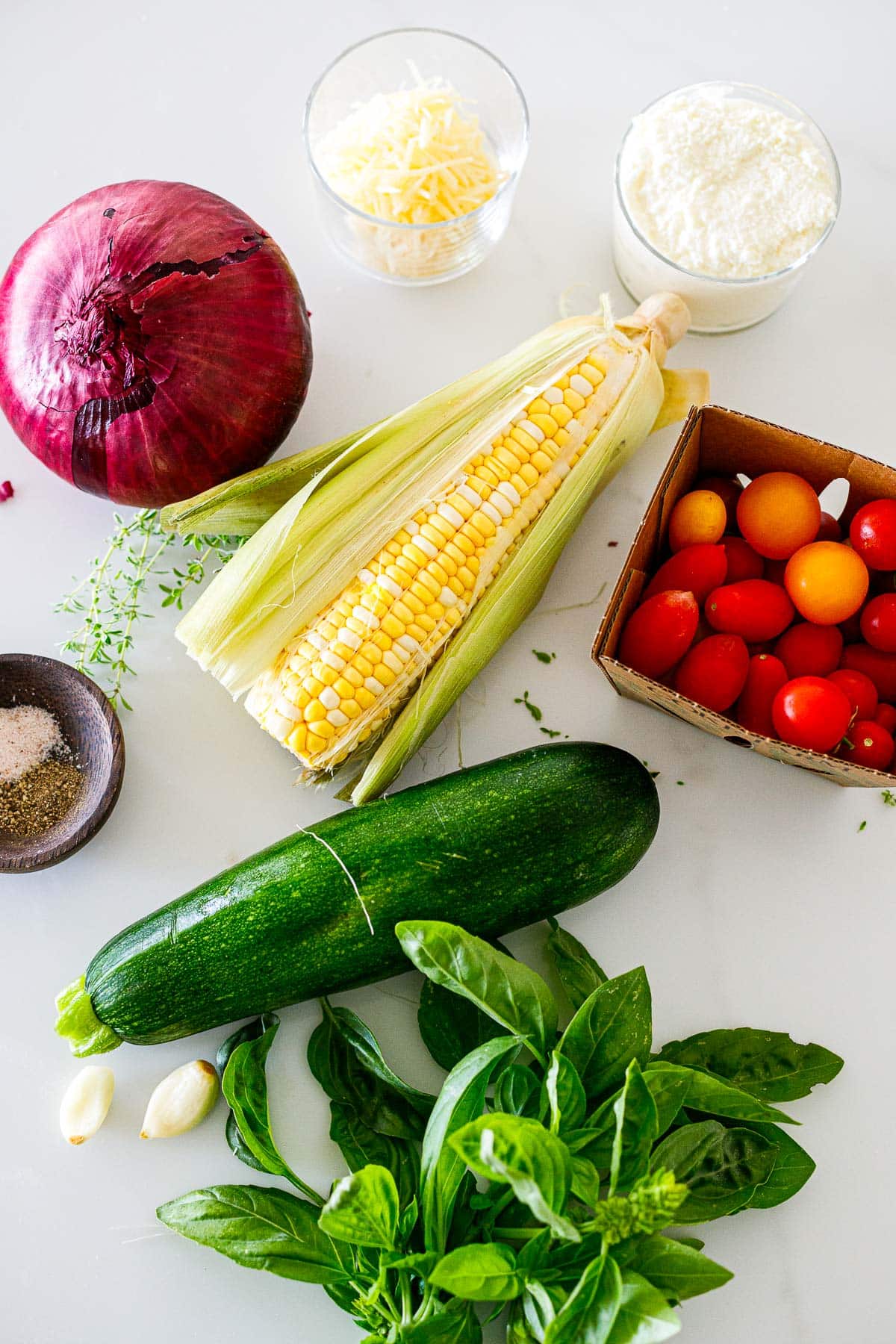 veggies laid out for savory summer galette filling- cherry tomatoes, zucchini basil, garlic, corn, and onion with jars of ricotta and parmesan.