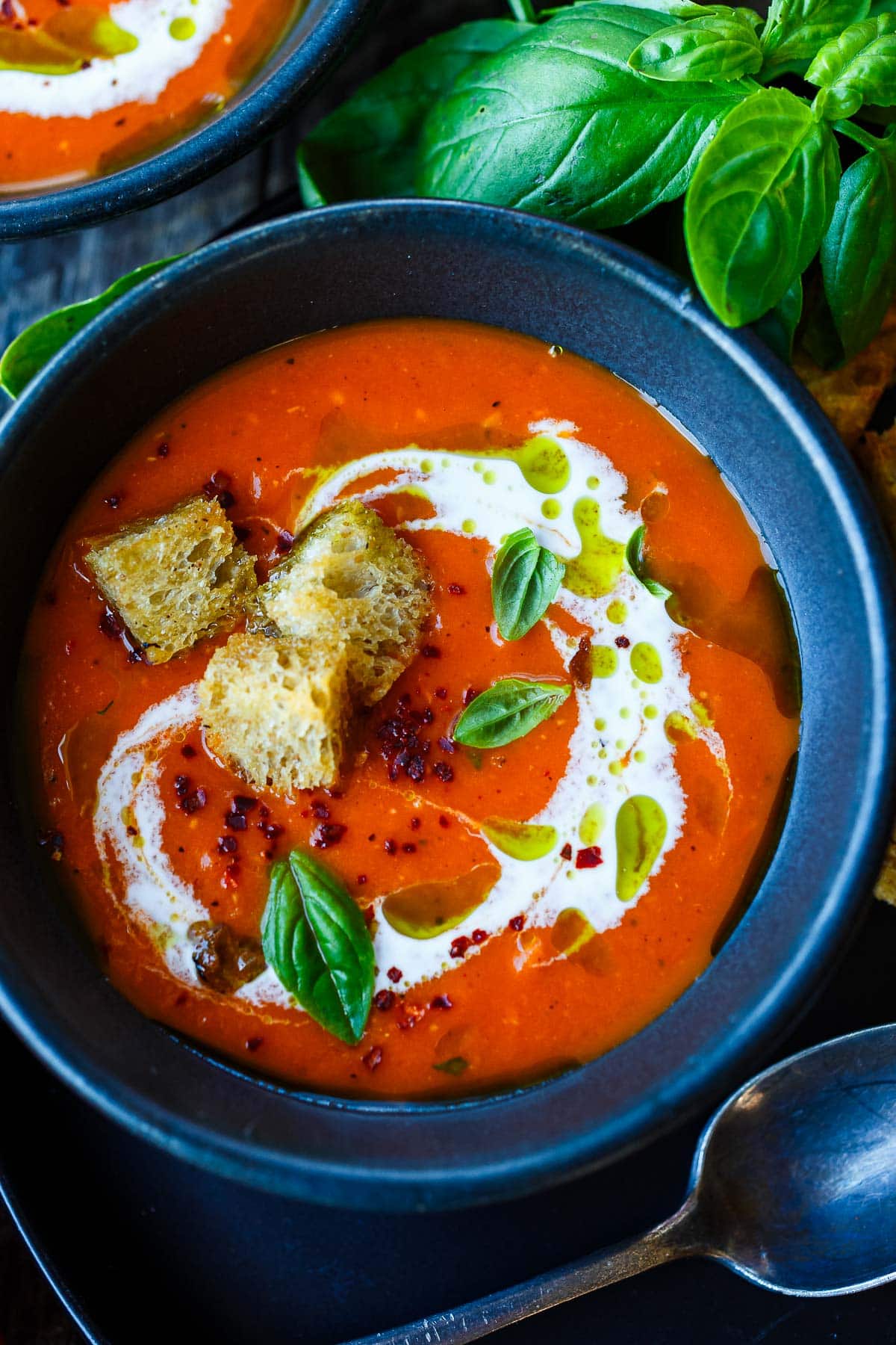 bowl of roasted red pepper and tomato soup with swirl of cream, drizzle of olive oil, fresh basil, and croutons. 