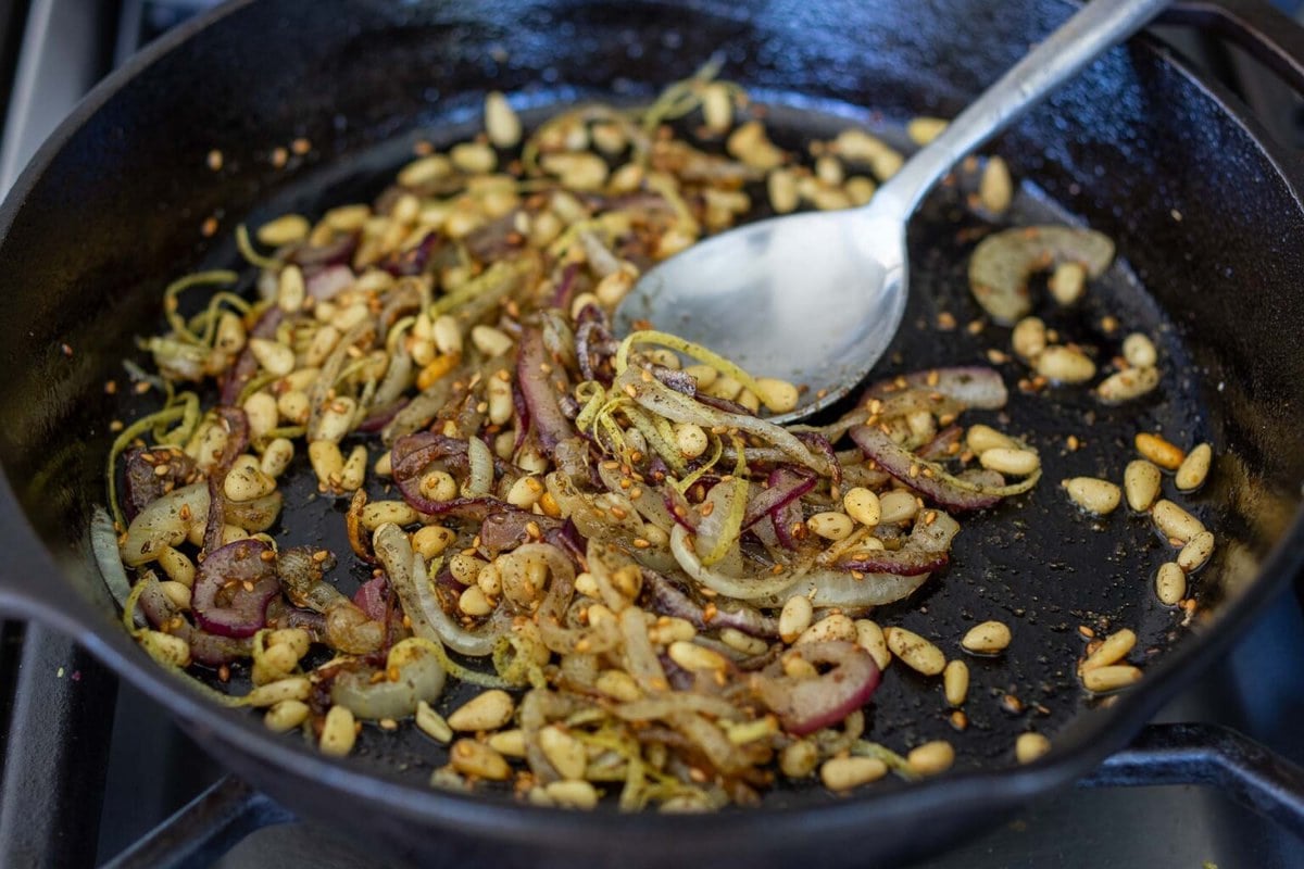 cast iron skillet with sauteed shallots, toasted pine nuts, lemon zest, and zaatar spice, stirred with metal spoon.