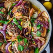 sumac chicken in a dutch oven with onions and sliced potatoes, topped with fresh herbs and lemon.