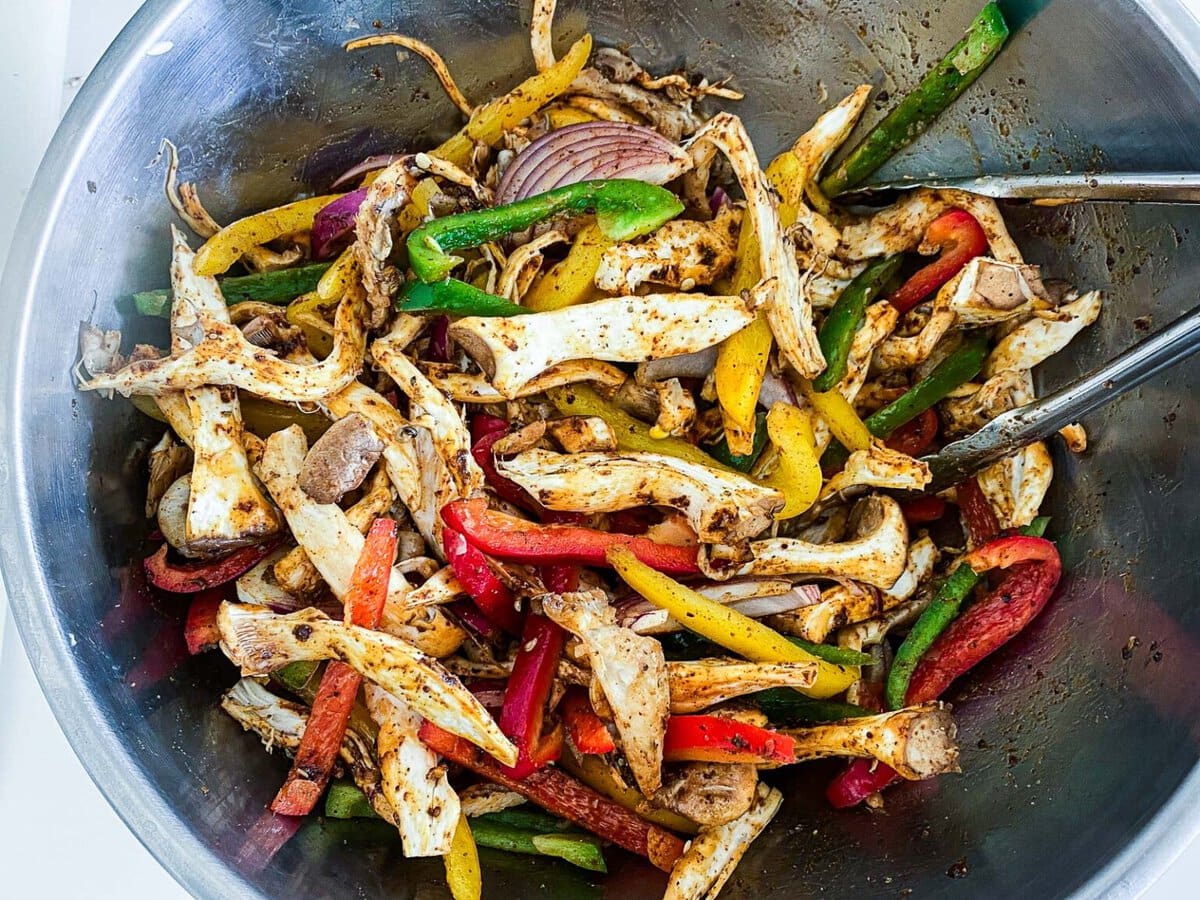mixing bowl and tongs with sliced fajitas veggies tossed in marinade.