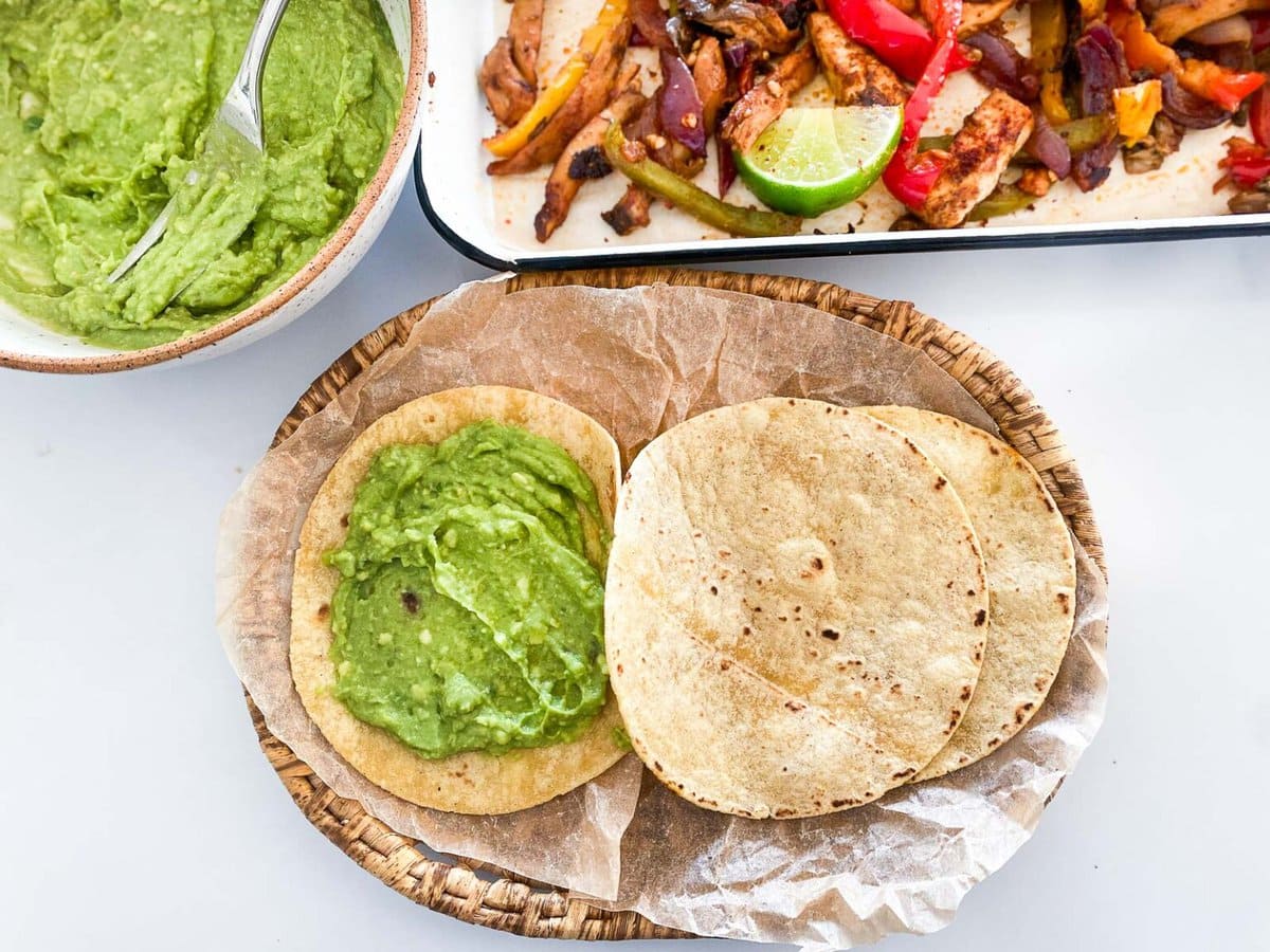 tortillas on parchment with one coated in avocado mashed next to bowl of avocado mash and pan of fajita veggies.