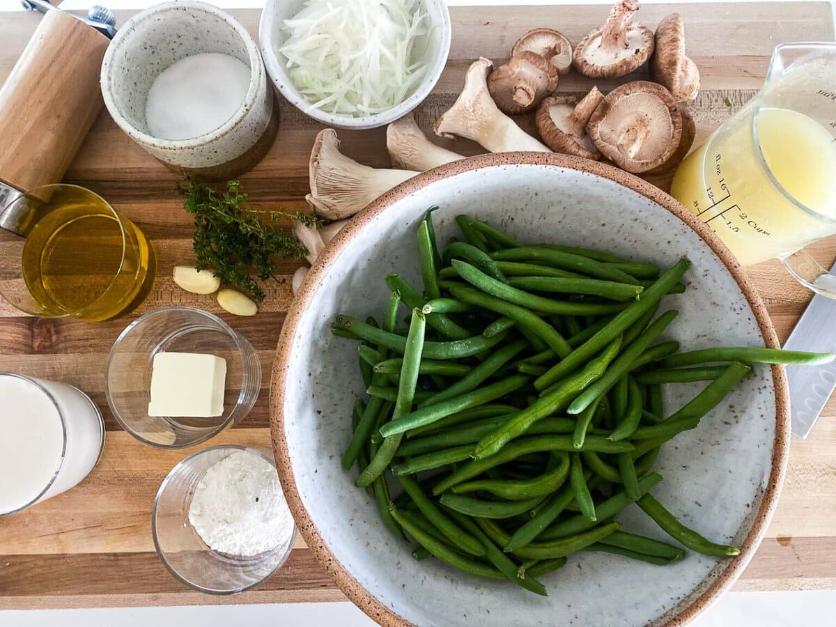 ingredients for green bean casserole - bowl of green beans, surrounded by jars of ingredients, garlic cloves, thyme, and mushrooms. 