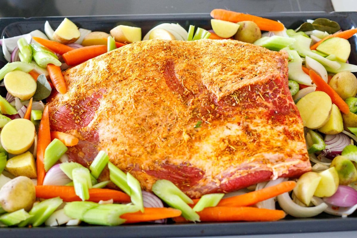uncooked brisket seasoned and on pan surrounded by vegetables.