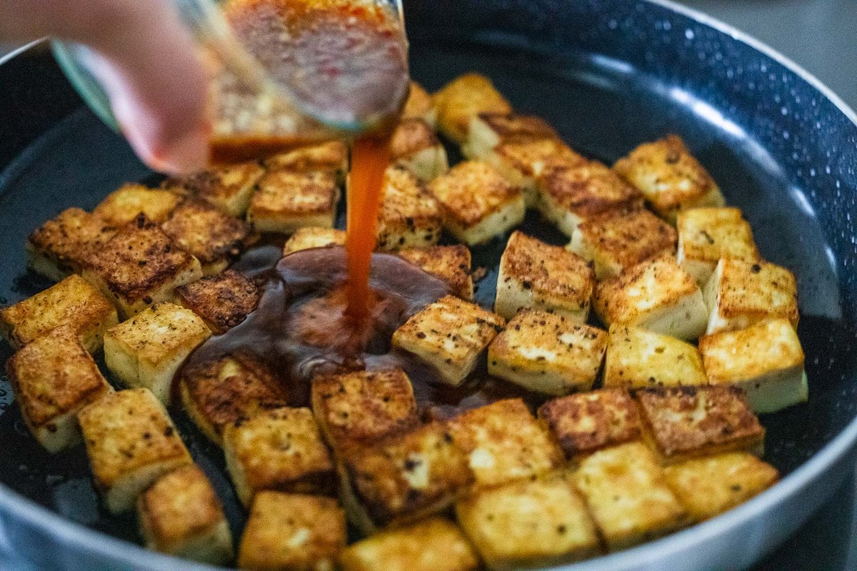 hand pouring sticky glaze over pan-fried tofu cubes.