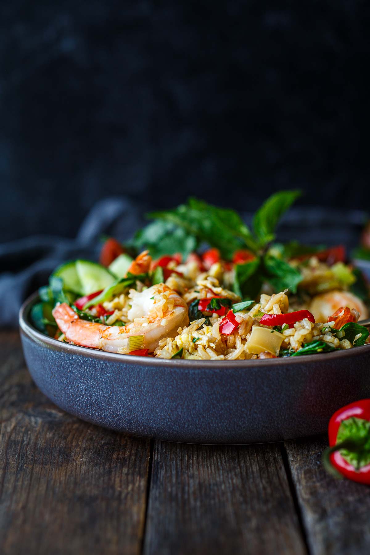 bowl of thai fried rice with shrimp served with a spoon, garnished with fresh herbs, sliced chilies, tomatoes, and cucumbers.
