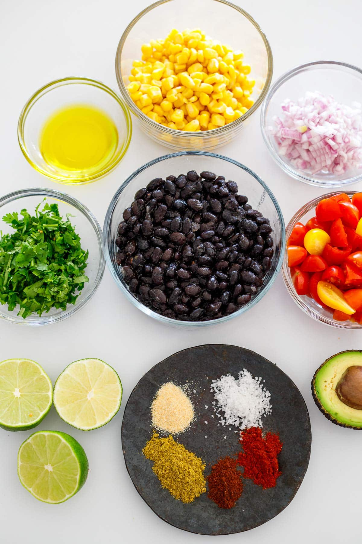 ingredients for black bean salsa arranged neatly in bowls - corn, onions, cherry tomatoes, black beans, olive oil, cilantro, avocado, limes, and spices.