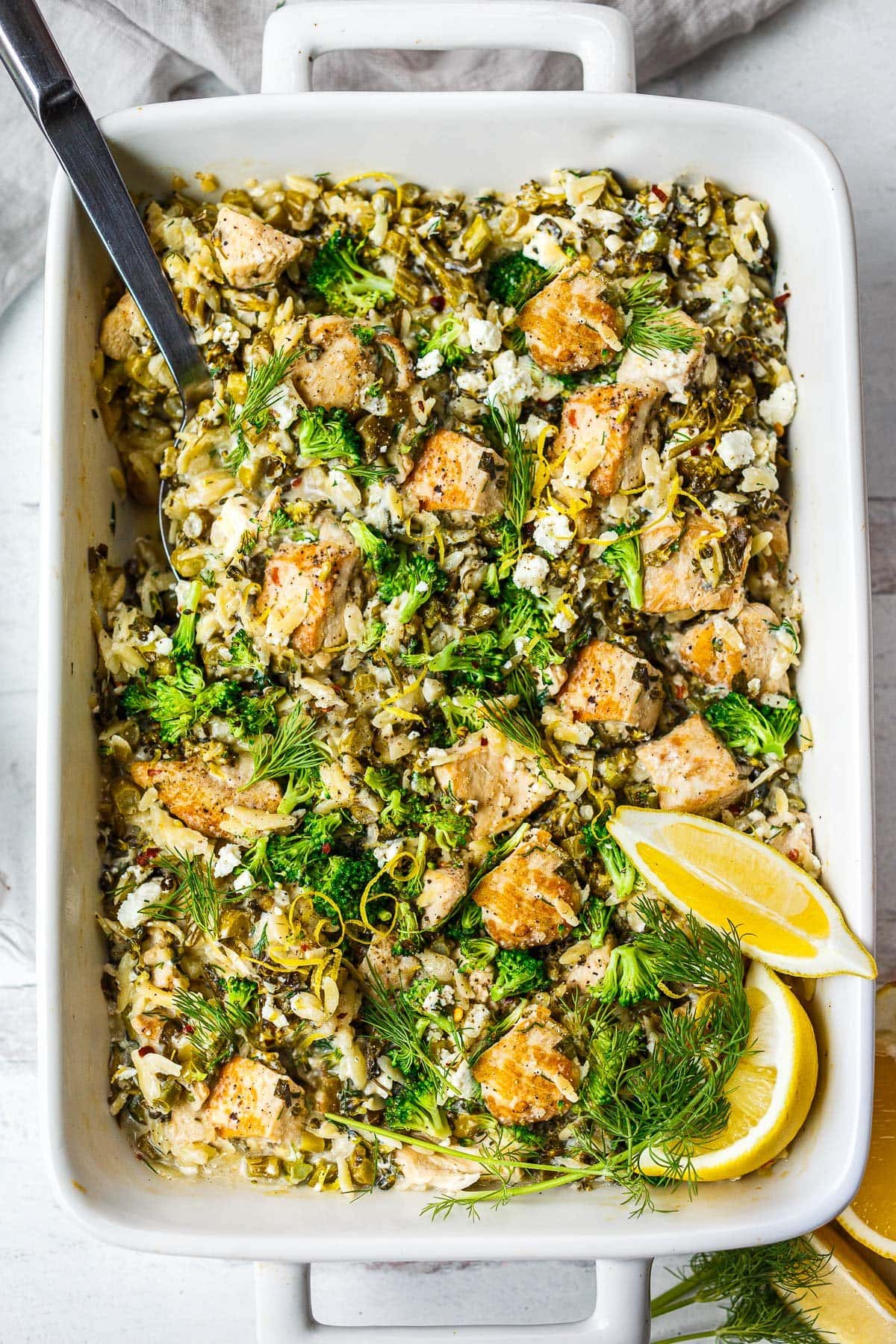 white ceramic baking dish with silver serving spoon scooping chicken orzo bake with feta, broccoli, dill, lemon zest and wedges. 