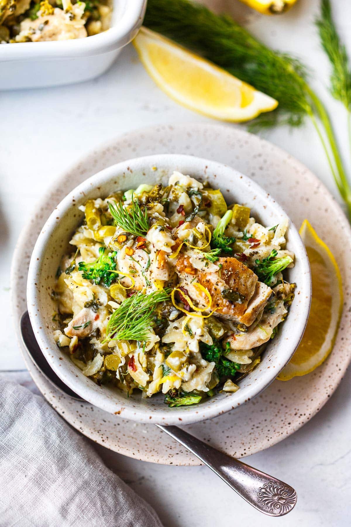 bowl of chicken orzo bake topped with fresh dill and lemon zest on small plate with lemon wedge and silver spoon.