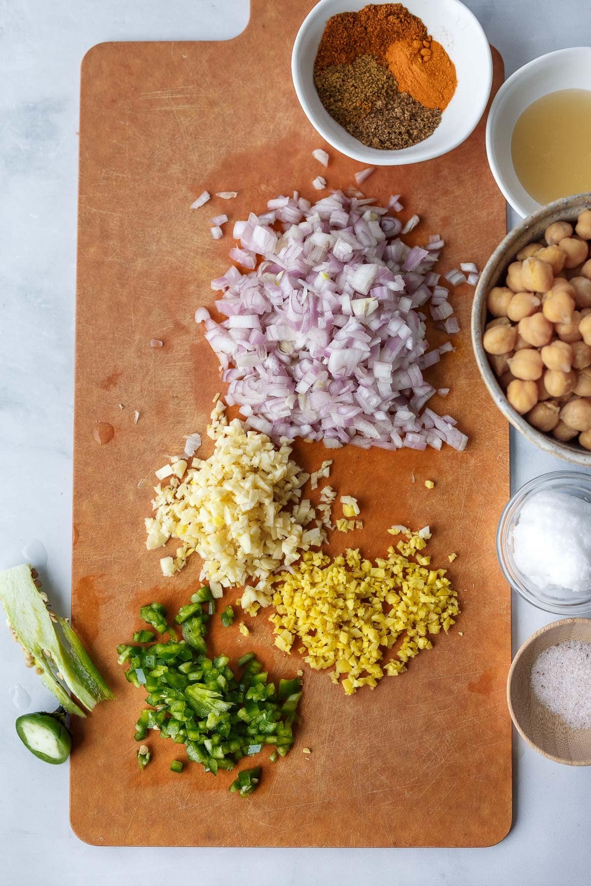cutting board with minced jalapeno, ginger, garlic, and shallot next to bowl of spices and other measured ingredients for chickpea curry.