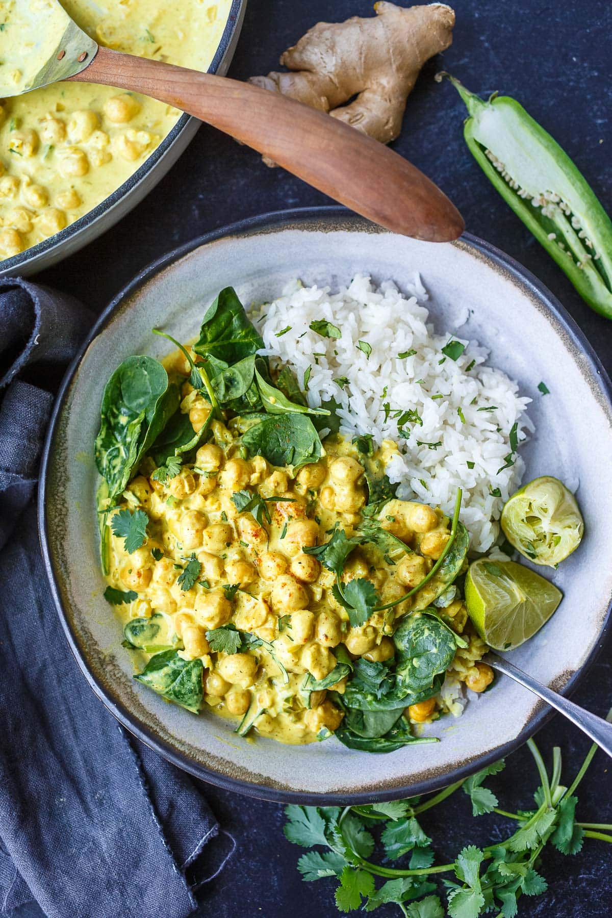 bowl of chickpea curry served with fresh spinach and jasmine rice, two lime wedges, and fresh cilantro.