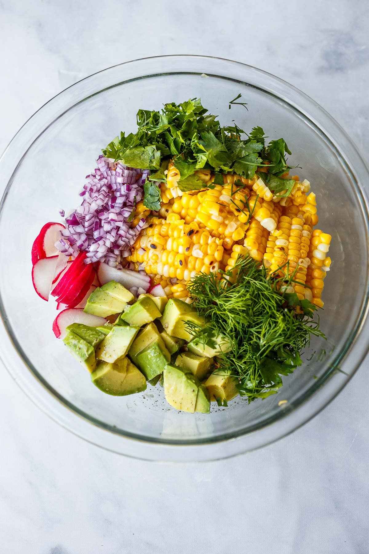 unassembled corn salad in glass mixing bowl - corn, dill, avocado, radish, red onion, cilantro.