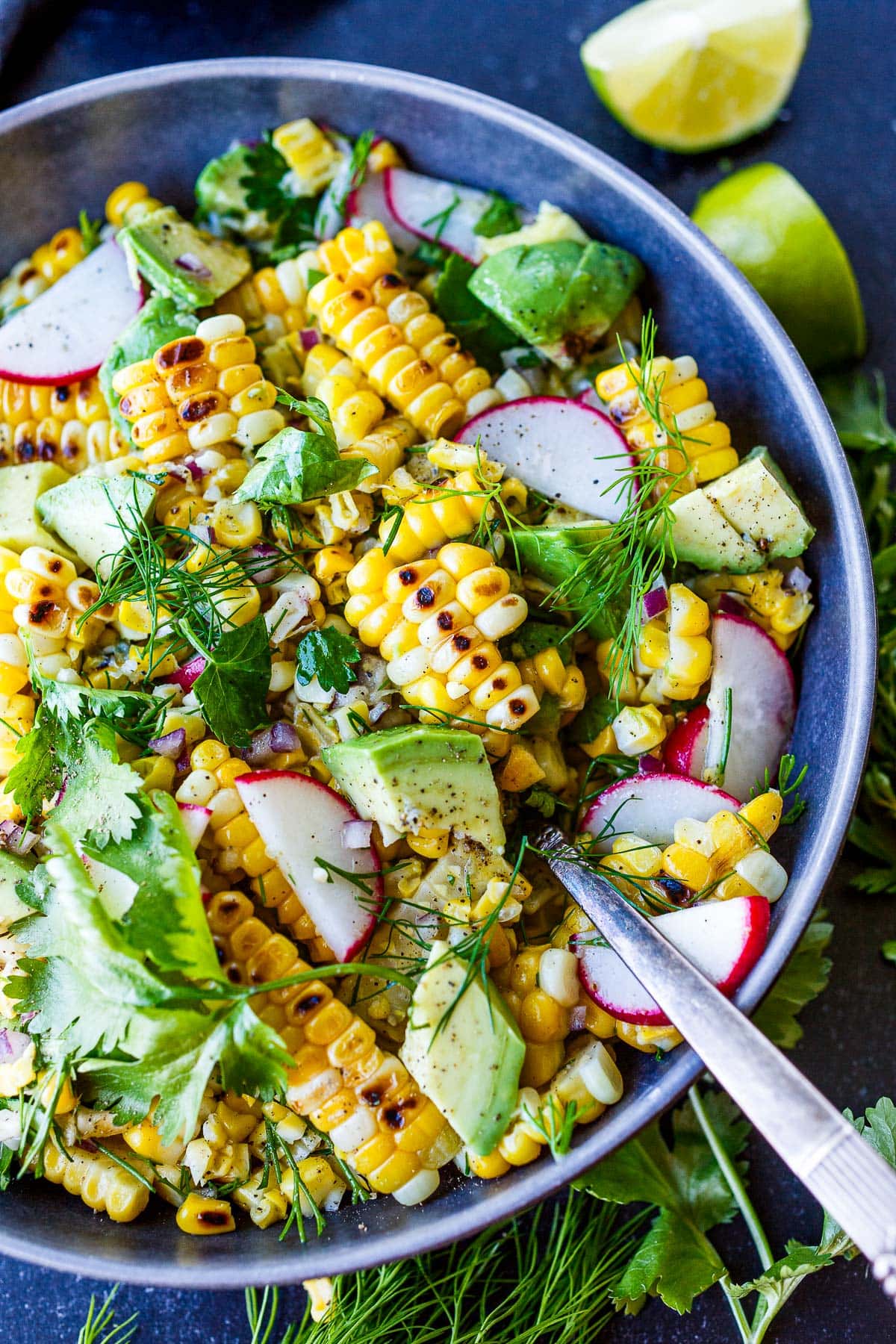 grilled corn salad in bowl with spoon with avocado, radish, dill, cilantro, and minced red onion.