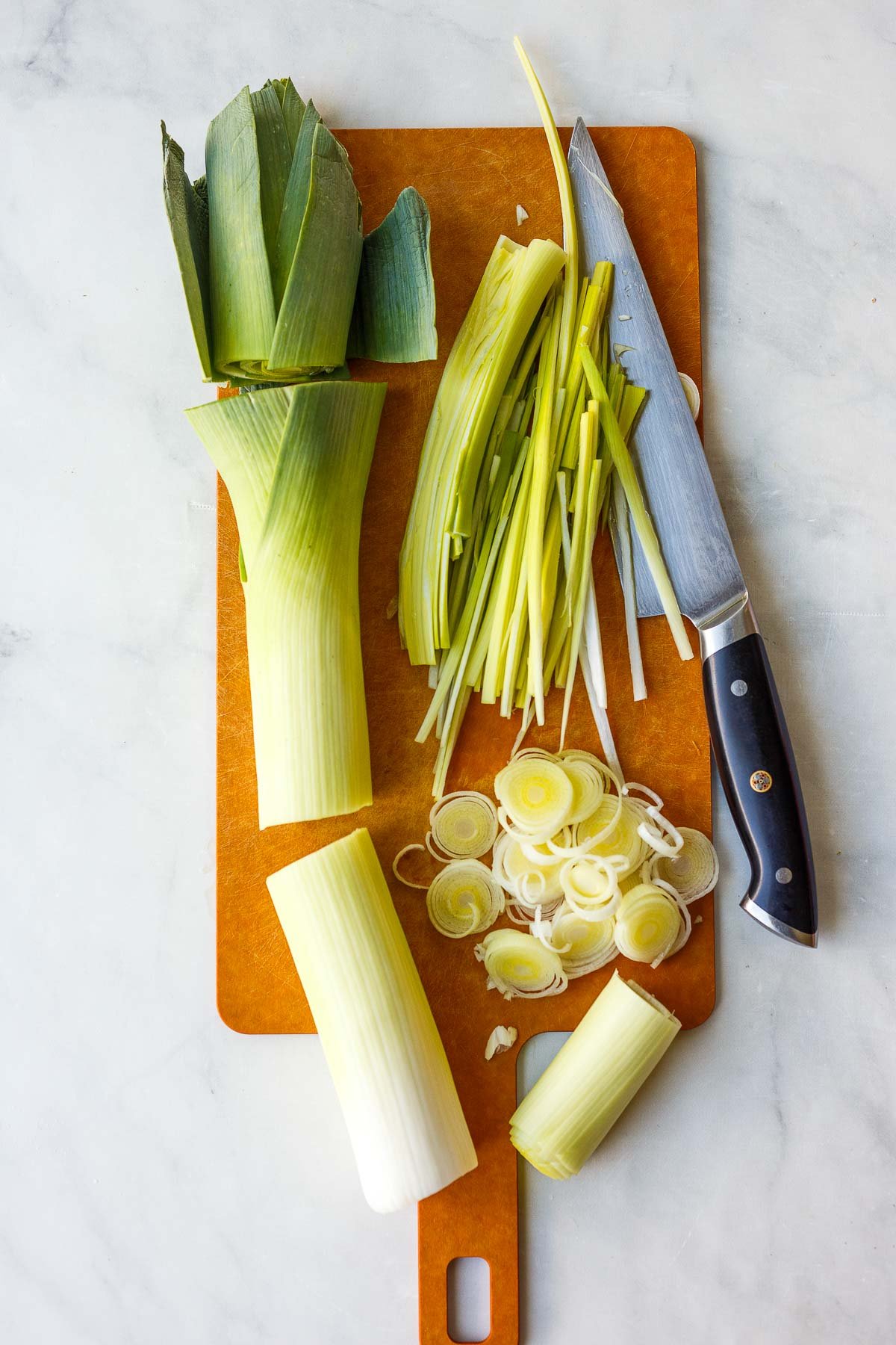 a large leek on a cutting board with the white section cut off and the rough green top cut off, next to the light green top thinly sliced in a pile, and the white part of the leek sliced into rounds in a pile on a cutting board with a sharp knife.