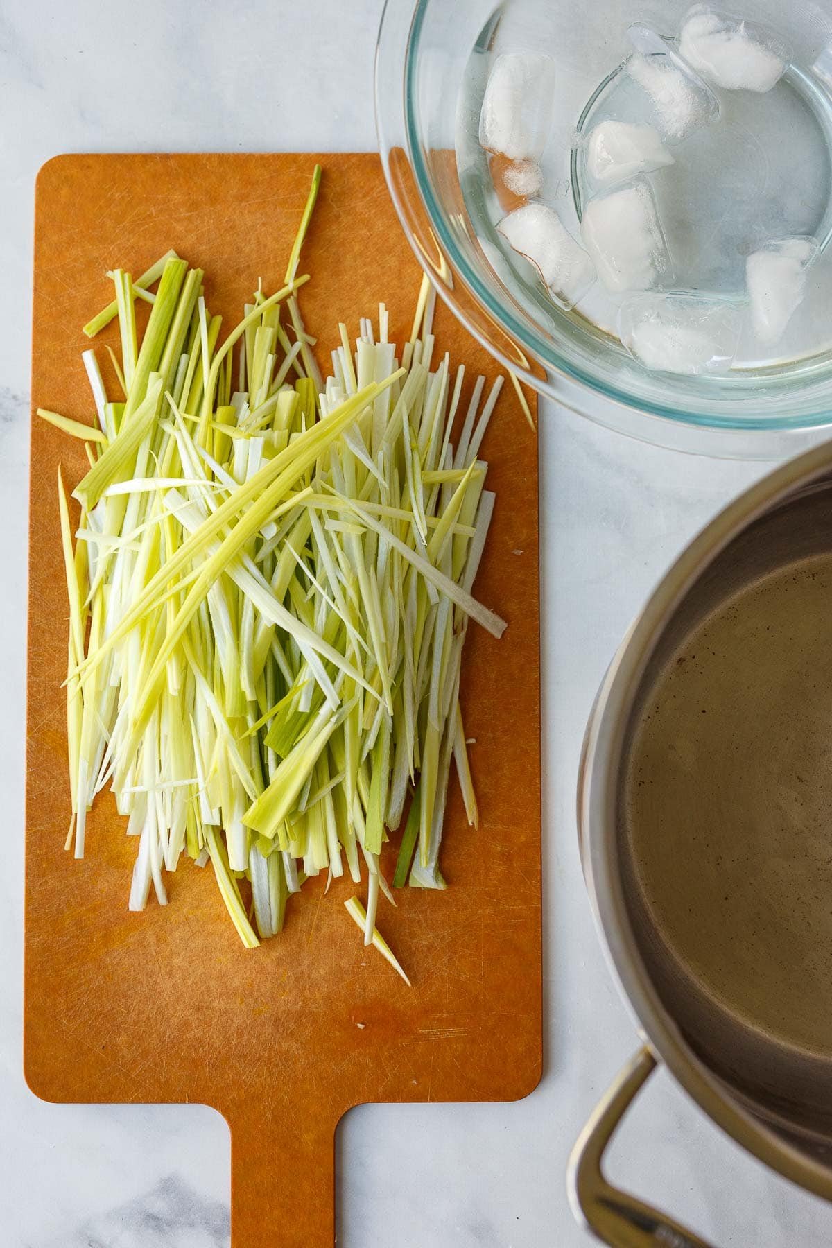 cutting board with light green part of leeks thinly sliced into strips next to a bowl of ice water.