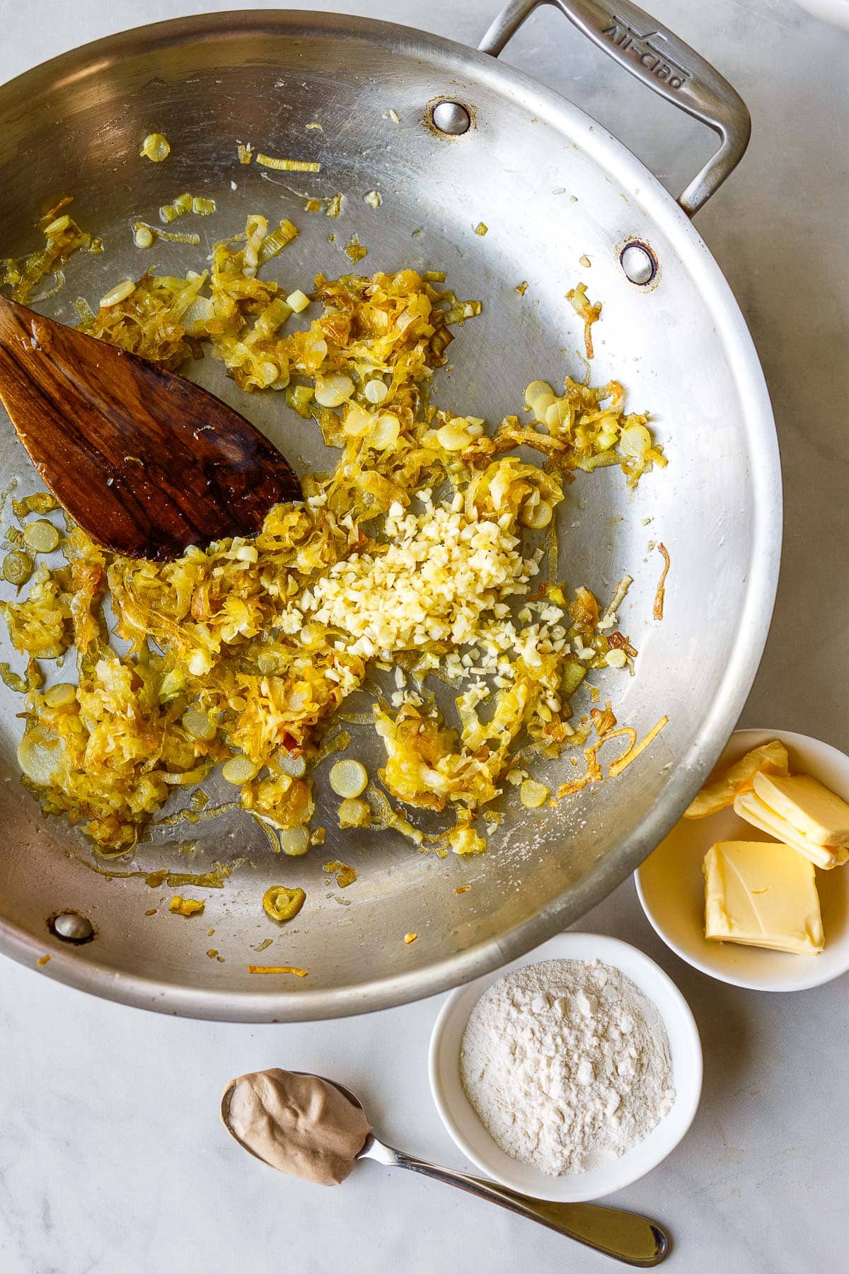 pan with caramelized leeks and minced garlic with wood spatula to stir next to small bowl of butter, flour, and a spoonful of dijon.