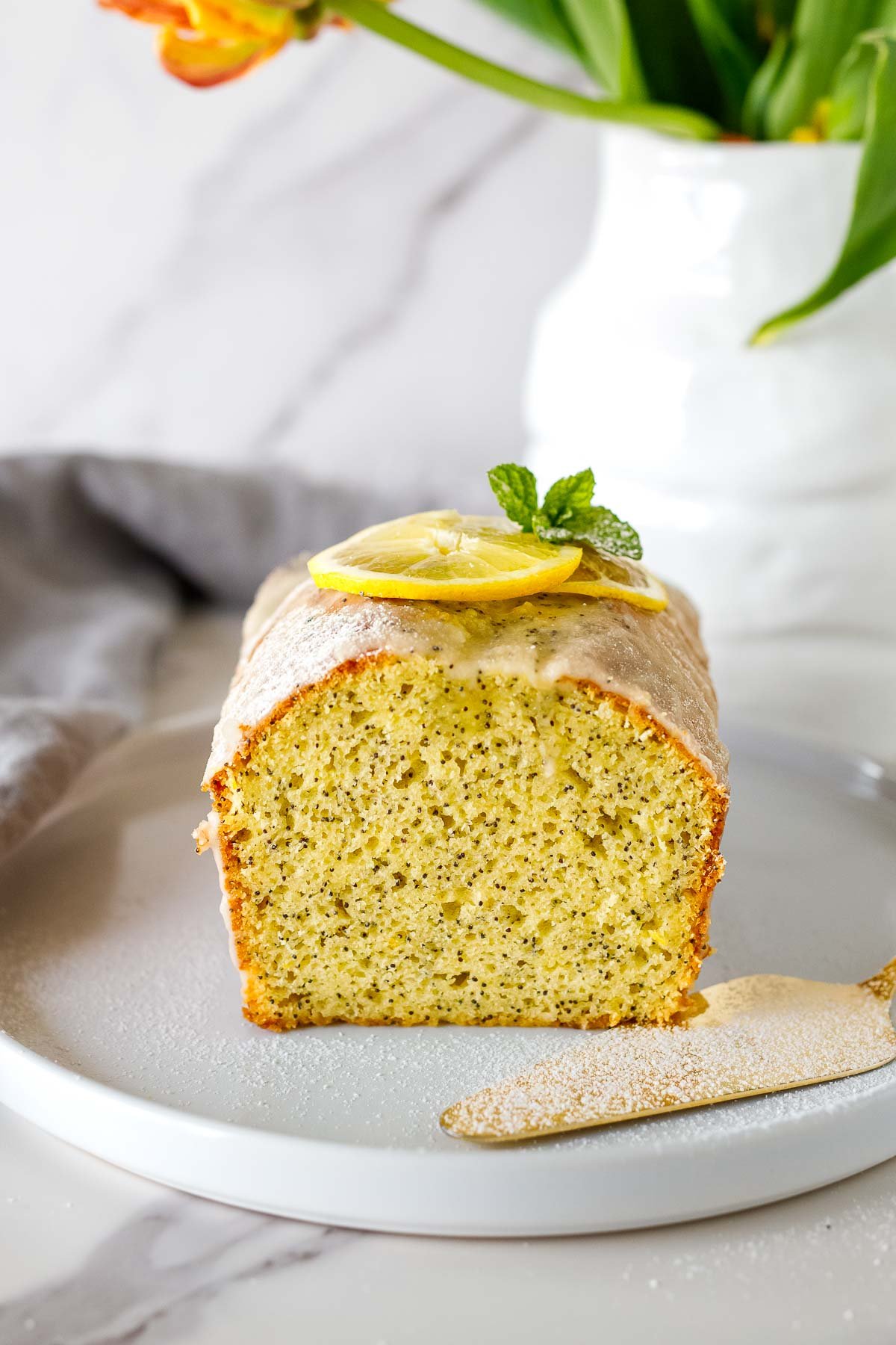 lemon poppy seed cake on plate garnished with lemon glaze, lemon slices, mint leaves, and powdered sugar.