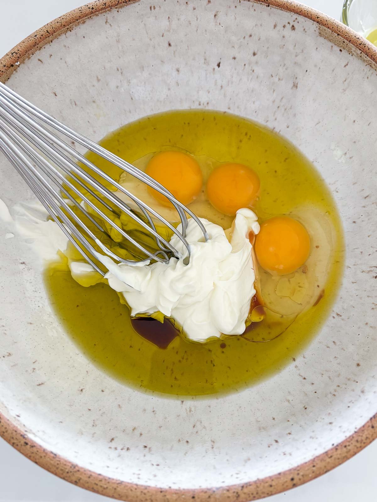 ceramic bowl with whisk stirring eggs, greek yogurt, olive oil, and vanilla.
