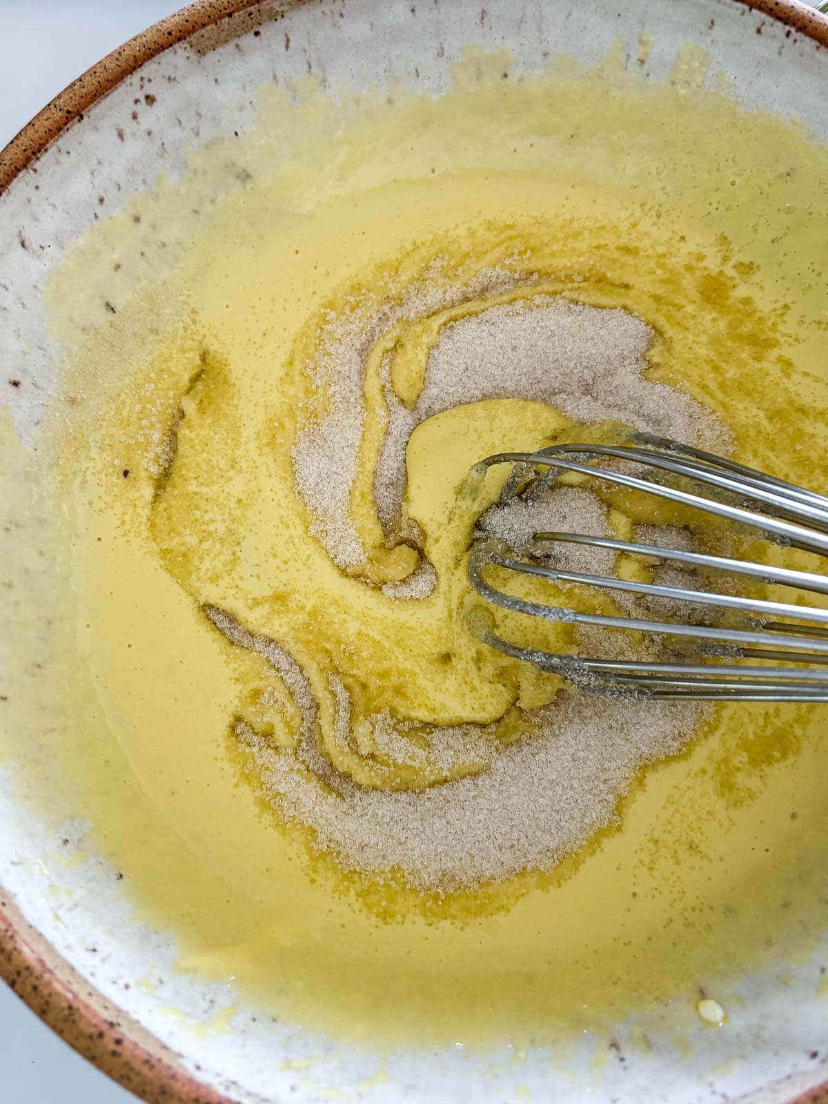 mixing bowl with whisk stirring sugar into wet ingredients for lemon poppy seed cake.