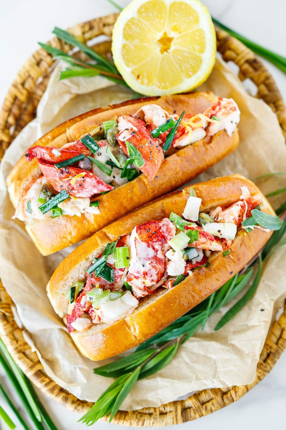 two lobster rolls in split-top buns topped with fresh herbs on parchment with a lemon half.