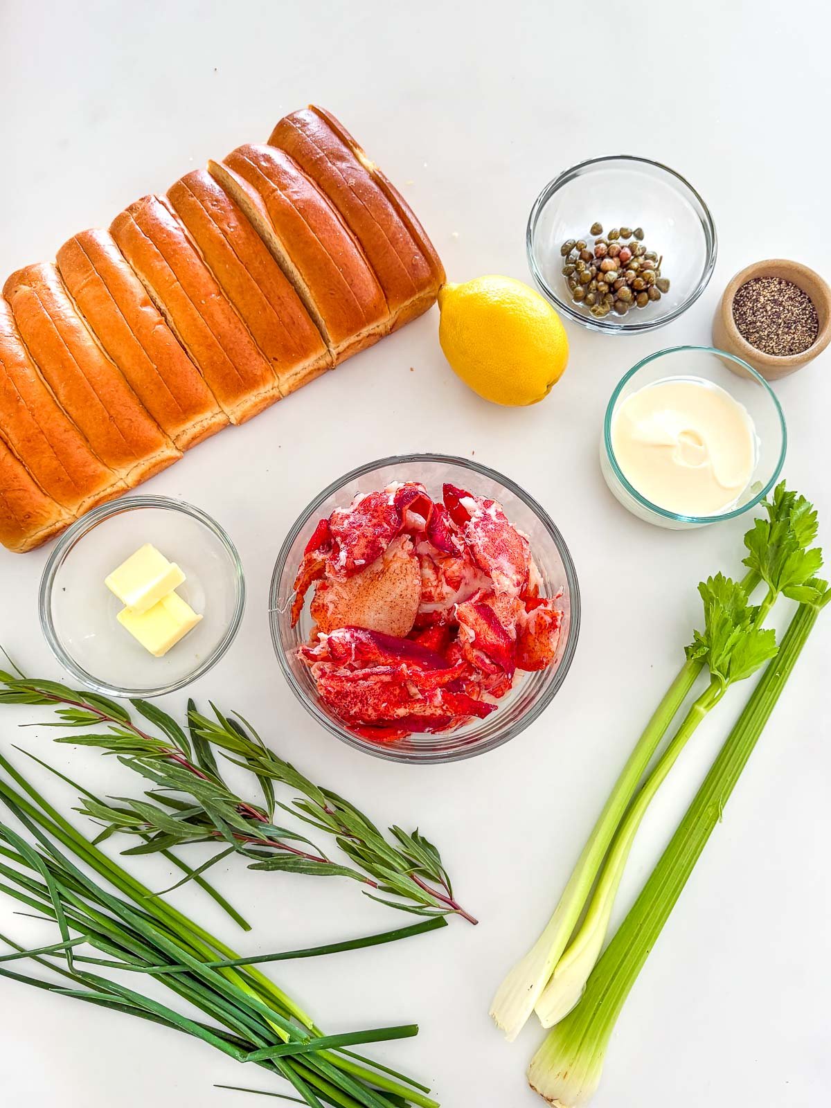 ingredients for lobster rolls arranged neatly - split-top buns, capers, lemon, mayonnaise, pepper, celery, lobster, butter, and herbs.