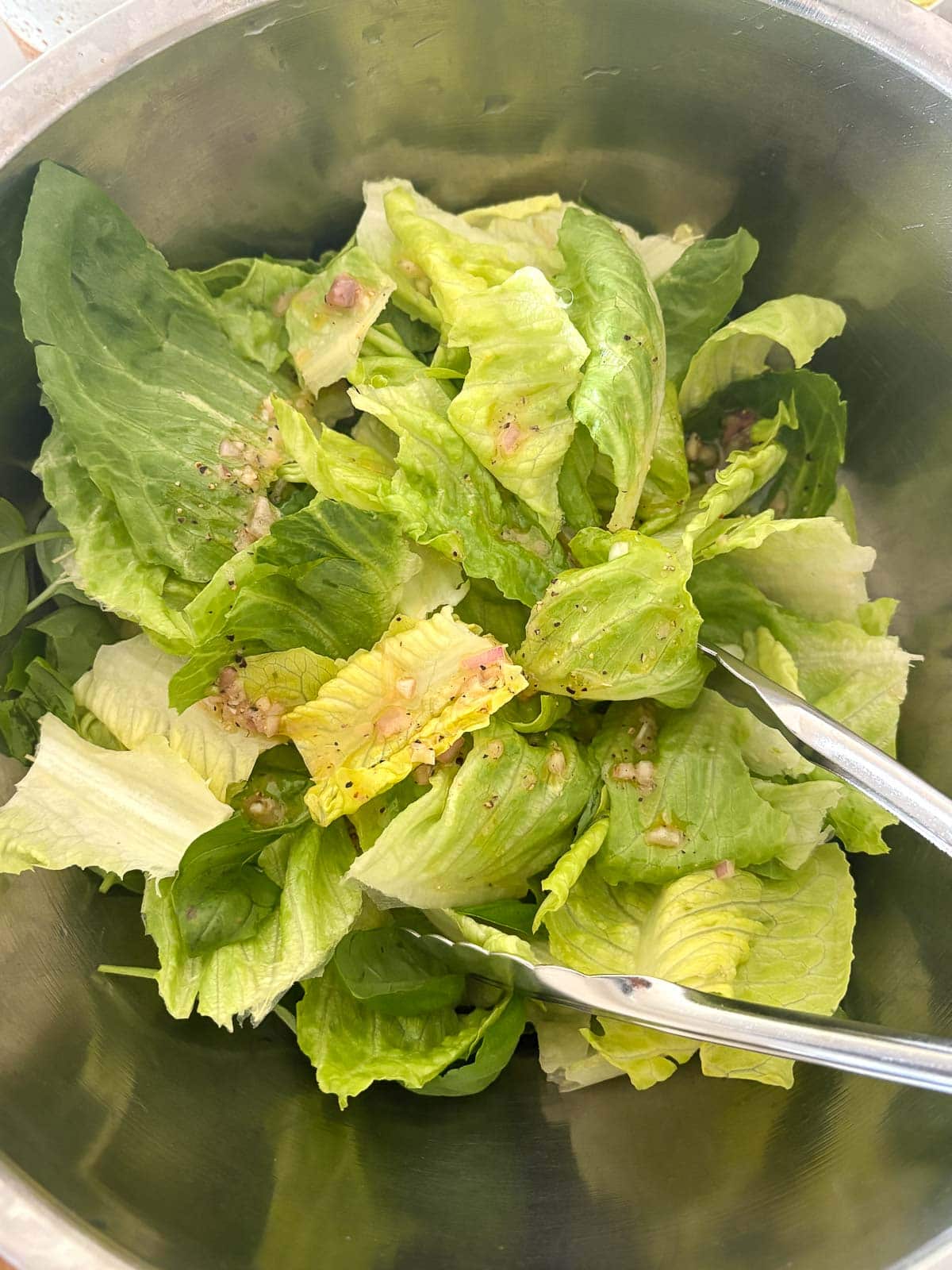 metal mixing bowl with crisp greens and tongs tossing lettuce in dressing.