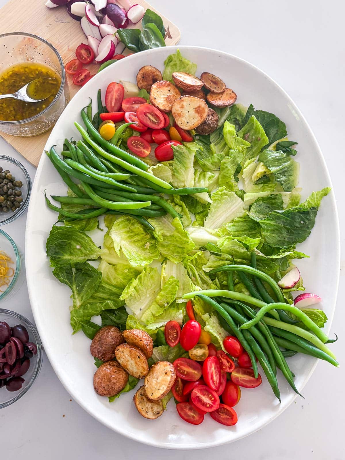 crisp greens, roasted potatoes, cherry tomatoes, and green beans arranged neatly on large oval platter to make nicoise salad.