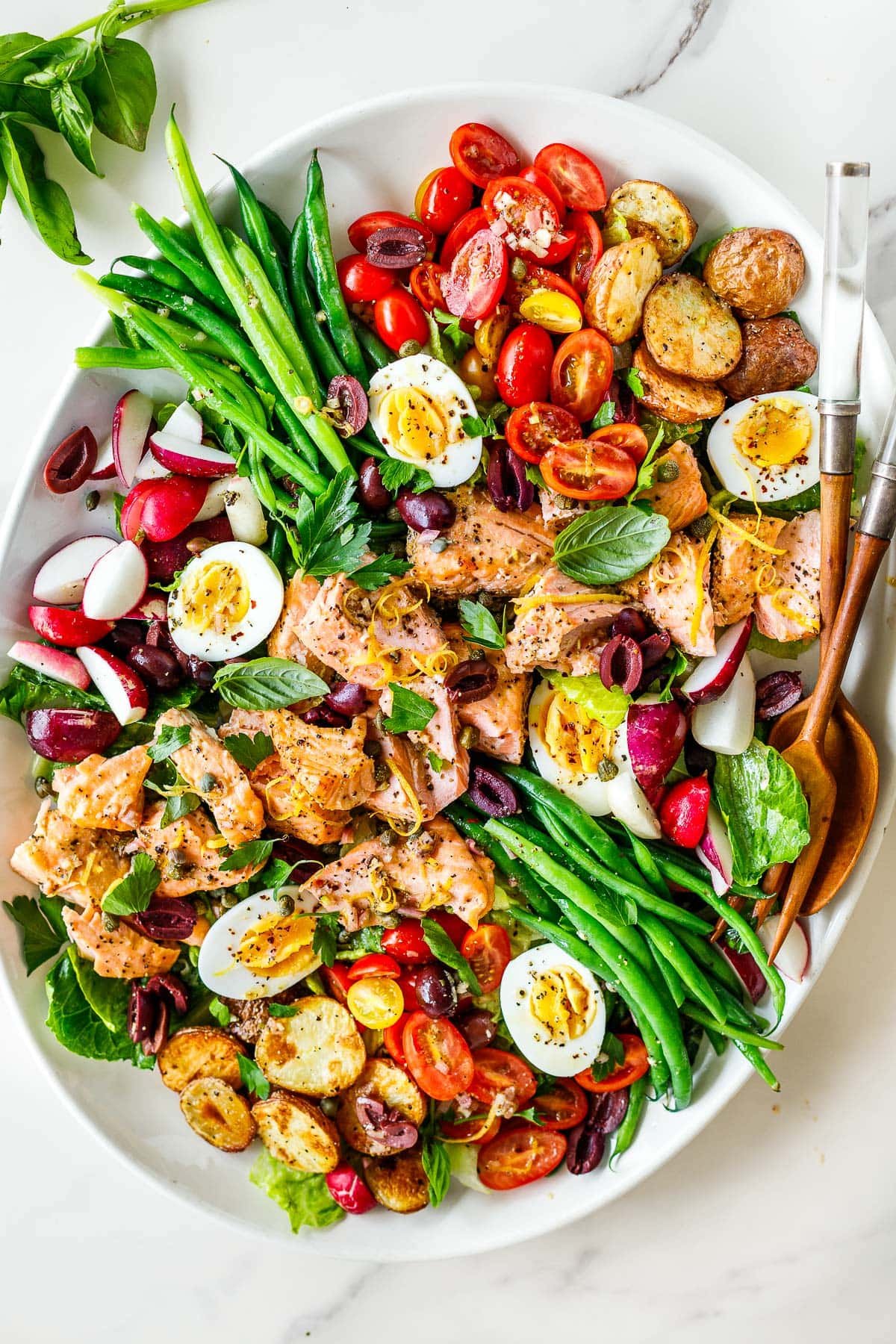 nicoise salad with baked salmon, boiled eggs, green beans, roasted baby potatoes, tomatoes, radishes, on large oval platter with wood serving utensils.
