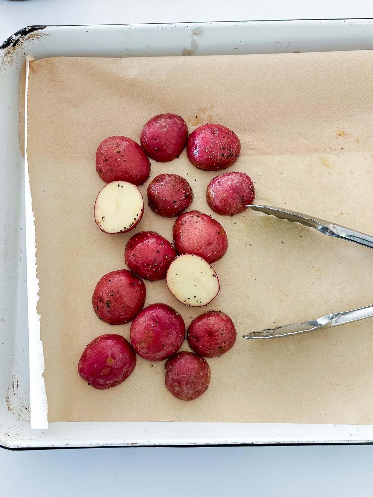 halved baby red potatoes seasoned on parchment lined baking sheet with tongs.