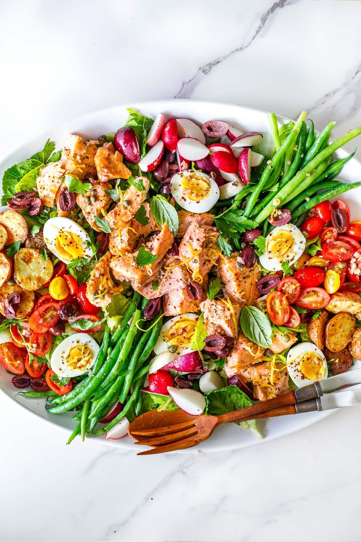 platter of nicoise salad with serving utensils, topped with baked salmon, boiled eggs, roasted potatoes, green beans, olives, capers, radishes, cherry tomatoes, with lemon zest.