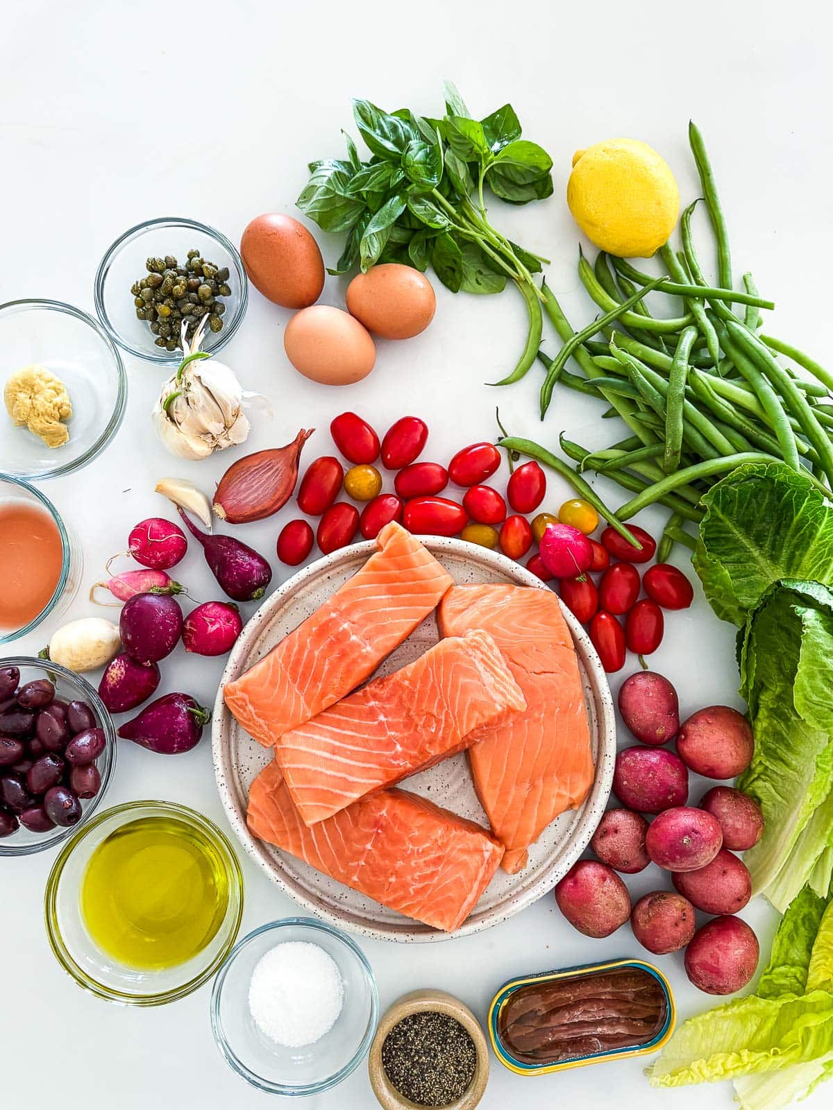 ingredients arranged to make nicoise salad - basil, lemon, green beans, lettuce, cherry tomatoes, baby potatoes, anchovies, pepper, salt, olive oil, olives, red wine vinegar, radishes, dijon, capers, garlic, shallot, eggs, all surrounding a plate of salmon filets.
