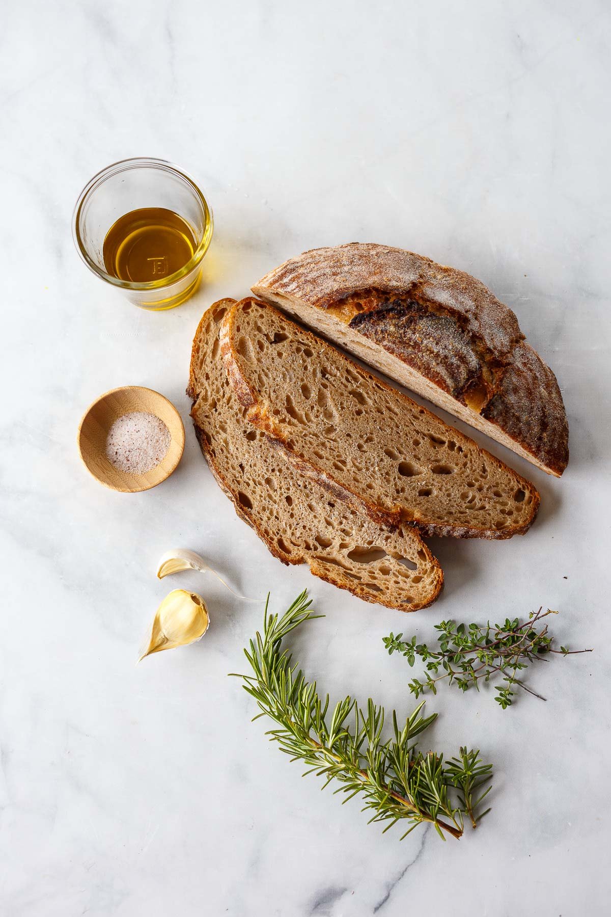 ingredients on marble counter to make pangrattato - olive oil, salt, sourdough bread, garlic, rosemary, thyme.