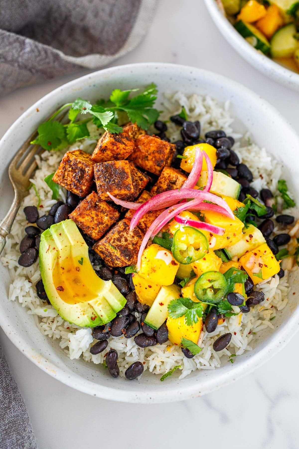 spicy jerk tofu bowl with jerk tofu over rice and beans topped with mango salad, pickled onions, and sliced avocado in bowl with fork.