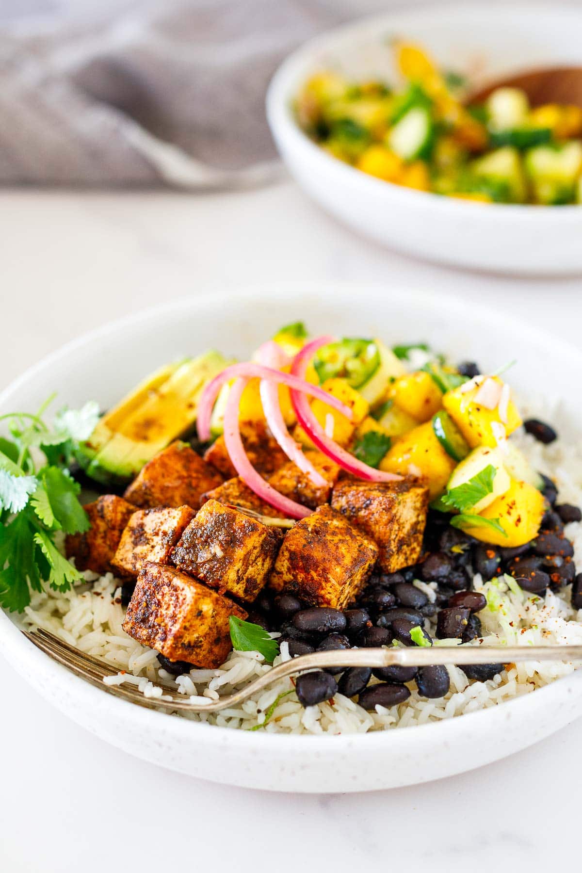 spicy jerk tofu bowl with rice, beans, mango salad, pickled onions, and cilantro in bowl with fork.